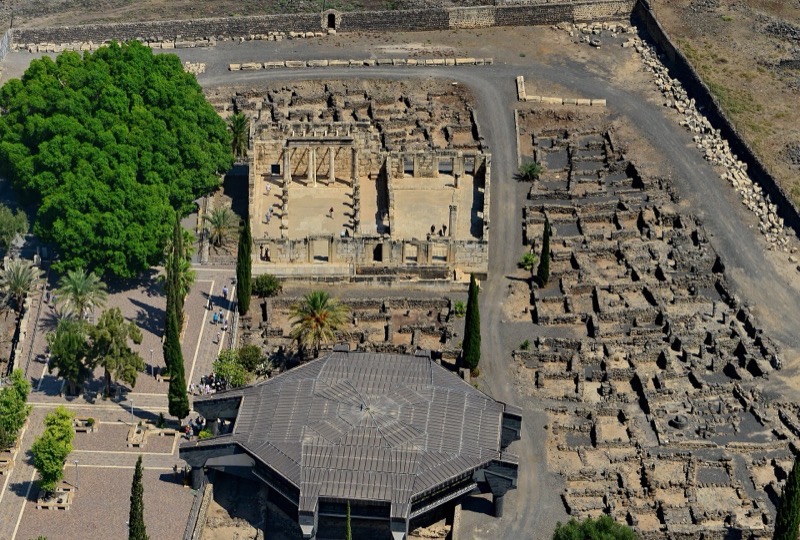 Aerial view of the excavated ruins at Tell Hum, identified with Capernaum: a white limestone synagogue among dark basalt house walls beside the Sea of Galilee